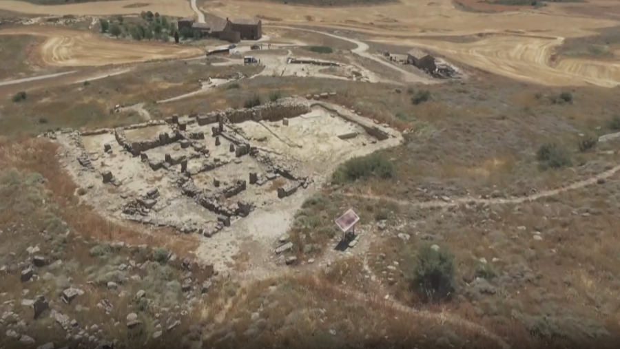 Vista aérea de ruinas romanas en España.  Se ven muros, cimientos y un cartel informativo cerca de un camino.  Campos rodean el sitio.