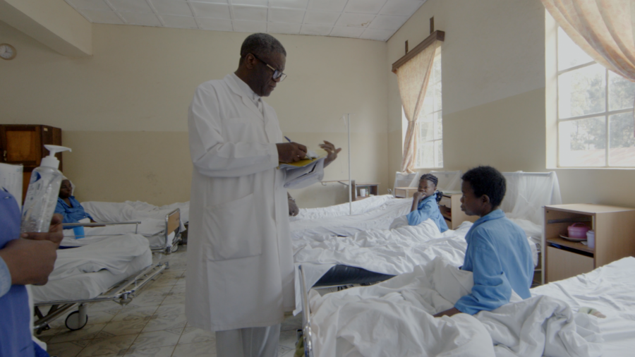 Médico tomando notas en un hospital con pacientes en camas, incluyendo niños. Personal con dispensador de gel. Posible escena del documental 'Semillas de Kivu'.