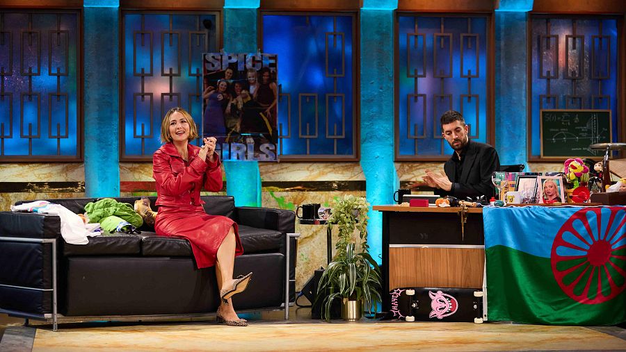 Entrevista en un programa de TV: mujer en traje rojo aplaudiendo frente a un presentador.  En el escritorio hay una bandera gitana, un monopatín y fotos.