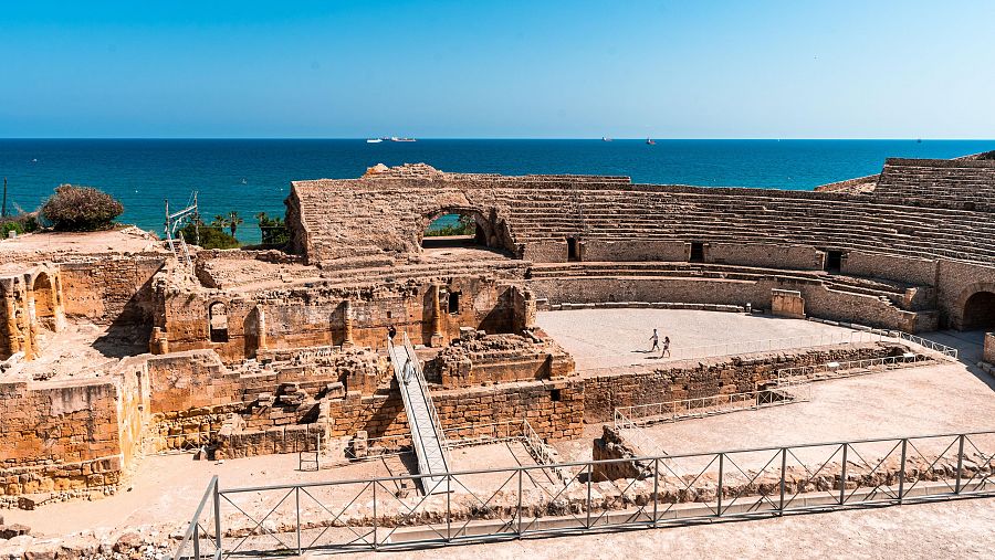 Anfiteatro romano en ruinas junto al mar. Dos personas caminan en la arena, barcos al fondo.  Restos de muros y gradas de piedra visibles.