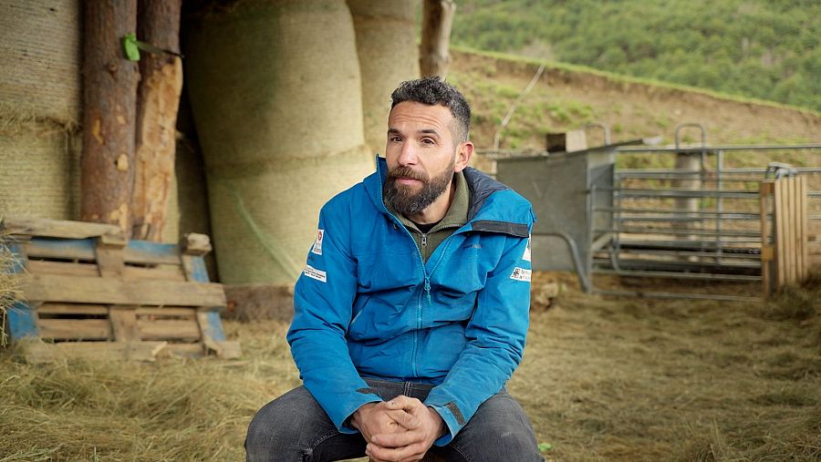 Hombre con barba y chaqueta azul sentado en un entorno rural, mirando hacia la izquierda.  Imagen promocional del programa 