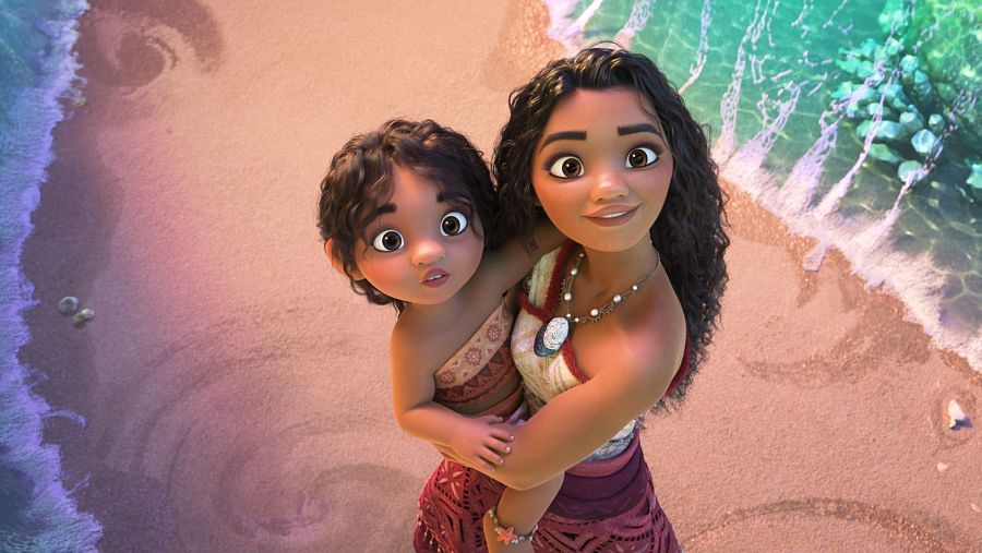 Vaiana y una niña, vestidas con atuendos polinesios, se abrazan en una playa.  Miran hacia arriba con expresiones amables.