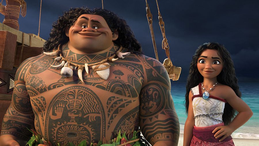 Fotograma de 'Vaiana 2': Vaiana y Maui, en una escena nocturna junto al mar y un barco. Maui luce tatuajes tribales y Vaiana viste un top blanco y falda roja.