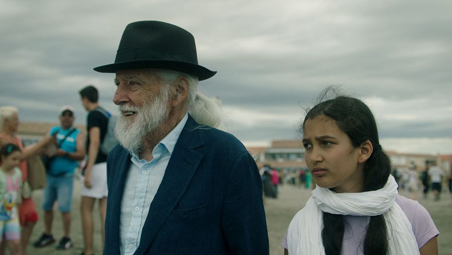 Un hombre mayor con barba blanca y una niña con pañuelo blanco miran a la derecha en una escena al aire libre de la película