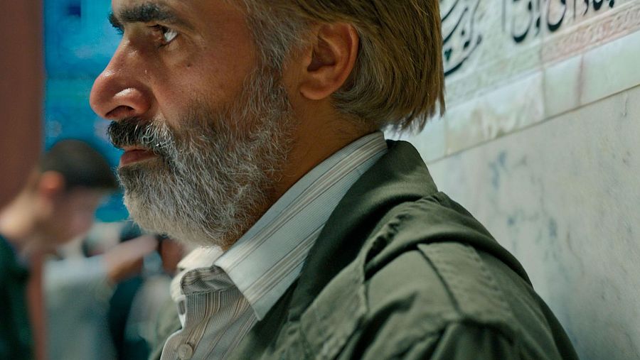 Hombre de mediana edad con barba y bigote canosos, camisa a rayas y chaqueta verde. Fondo borroso con escritura árabe o persa. Posiblemente relacionado con la película 'Araña Sagrada'.