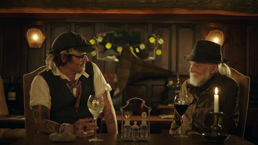 Charles Chaplin, con sombrero y barba, conversa con otro hombre en un bar de época.  Ambientes cálidos, copas de vino y ginebra en la mesa.