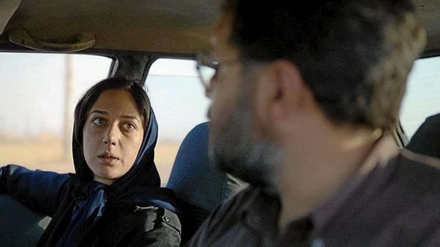 Mujer con pañuelo y hombre con gafas en un coche. Paisaje desértico visible por la ventana. Escena de la película 