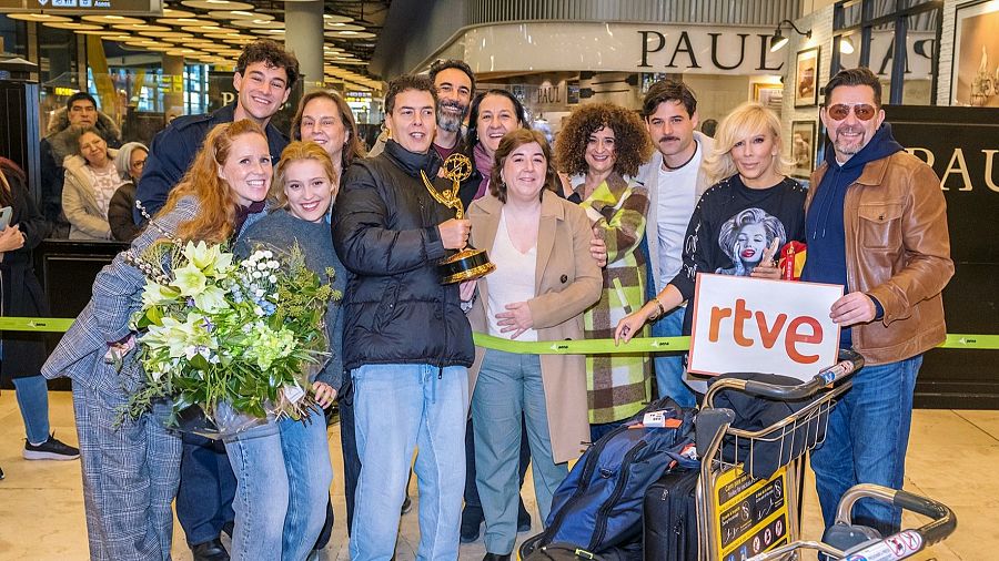 Equipo de 'La Promesa' celebra su Emmy en Barajas.  Posan con el trofeo y flores. Cartel de RTVE visible.