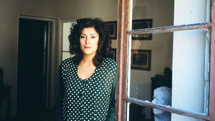 Retrato de Almudena Grandes, escritora española, con blusa verde con lunares blancos, junto a una ventana.  El texto 'Somos Cine' sugiere una posible entrevista o evento cinematográfico.