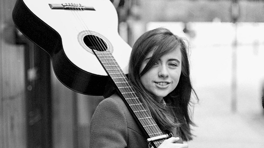 Mari Trini joven, en blanco y negro, con guitarra, sonriendo a cámara. Años 70. Fotografía callejera.