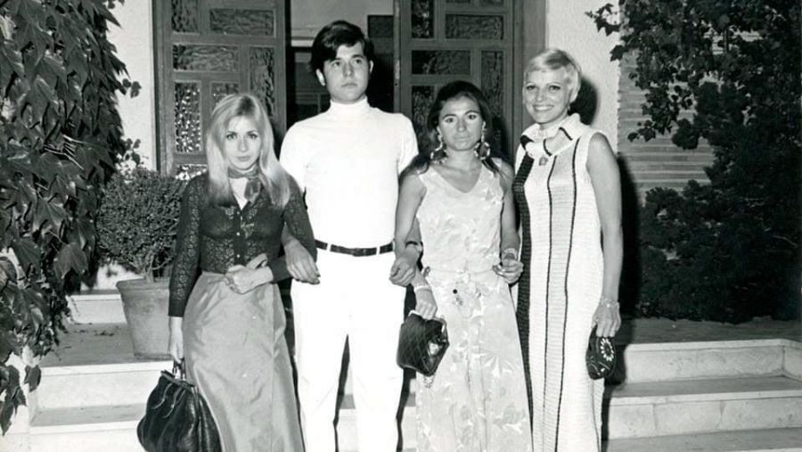 Cuatro personas vestidas para una ocasión especial posan en la entrada de una casa con plantas a ambos lados de la puerta.