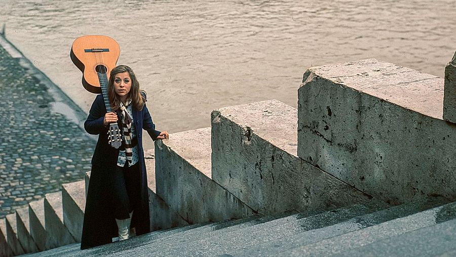 Mari Trini en París, bajando una escalera de piedra junto a un río con su guitarra. Viste abrigo oscuro y botas claras.