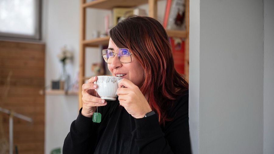 Elena Crespi bebe té, sonriendo. Lleva gafas y una blusa negra.  Fondo con libros y texto: 