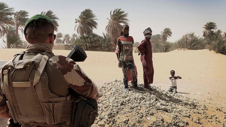 Soldado en Chad interactuando con tres niños.  Uniforme de camuflaje, chaleco antibalas visibles. Fondo desértico.