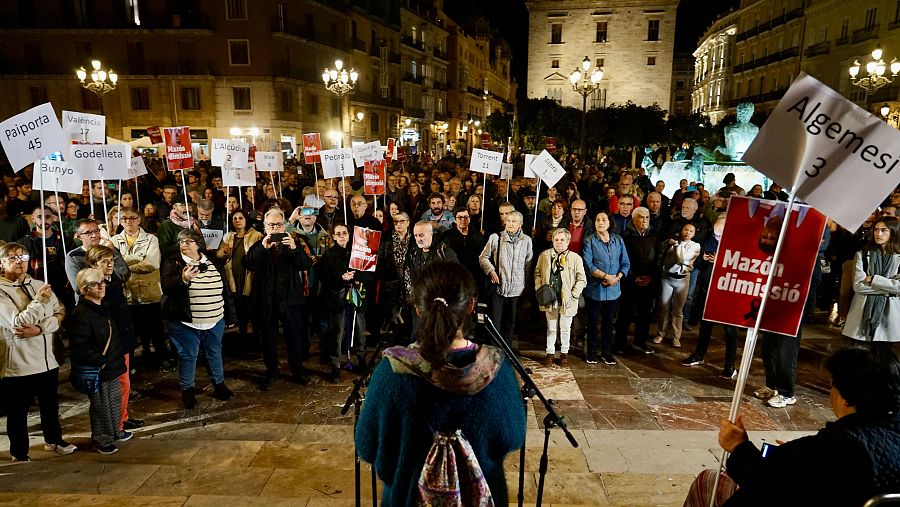 Protesta nocturna en la Plaza de la Virgen, Valencia, contra Carlos Mazón.  Carteles con nombres de pueblos y peticiones de dimisión.  Orador con micrófono.