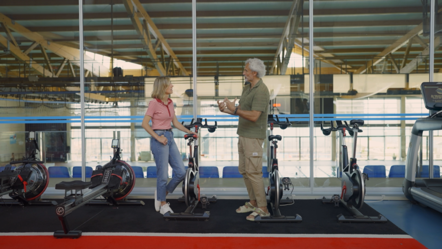 Antonio Jorge Larruy conversa con una mujer en un gimnasio con bicicletas estáticas y máquinas de remo.  Él gesticula mientras habla.