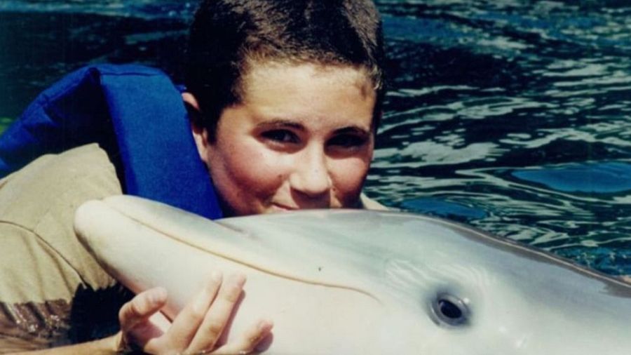 Mujer con chaleco salvavidas azul abraza a un delfín gris claro en aguas azules oscuras. Interacción cercana y amistosa.
