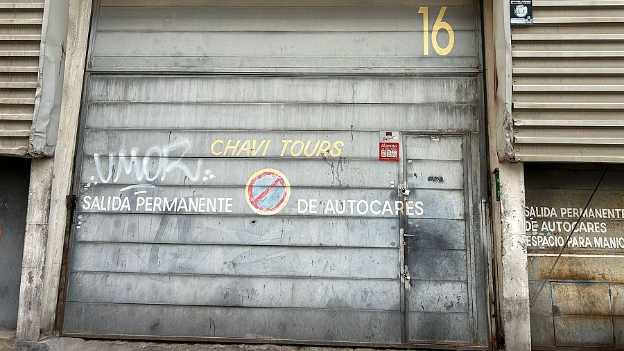 Fachada de Chavi Tours con puerta metálica gris, letreros 'SALIDA PERMANENTE DE AUTOCARES' y grafitis. Se ve el número 16 y una segunda puerta más pequeña.