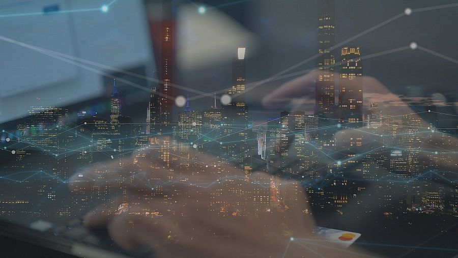 Ciudad nocturna superpuesta a manos usando una tablet y una tarjeta de crédito, representando conexiones digitales y posibles transacciones.