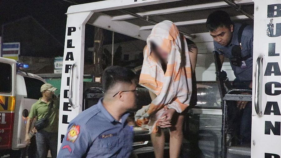 Un individuo con la cara tapada con una tela naranja y blanca es detenido por dos policías y subido a un vehículo policial con la inscripción 