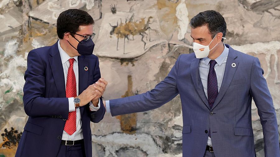 Dos hombres en trajes formales se estrechan la mano; uno lleva una mascarilla blanca con la bandera de España, el otro una negra.  Imagen formal, probablemente un saludo profesional.