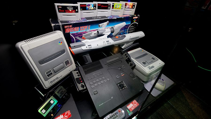 Consolas y videojuegos retro, entre ellos una Super Nintendo con su pistola Scope 6 y cartuchos, una consola negra tipo ordenador y otros mandos y accesorios.  Pegatinas con el logo 