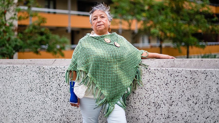 Mujer mayor con poncho verde y férula en la mano izquierda, apoyada en un muro. Fondo con edificio y vegetación.