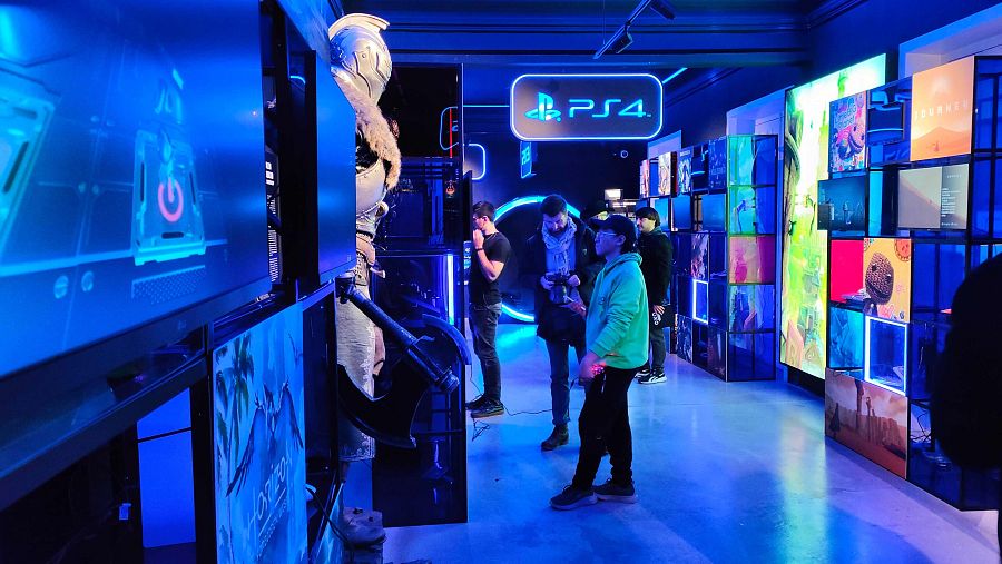 Exposición interactiva de Playstation con pantallas, figuras de videojuegos y objetos coleccionables. Iluminación azul futurista.  Visitantes exploran la muestra.