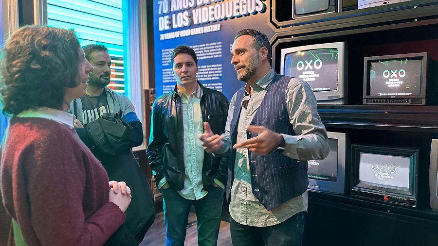 David Bustamante en una exposición de videojuegos, conversando con otras personas ante pantallas con el juego OXO. Se observa el cartel 
