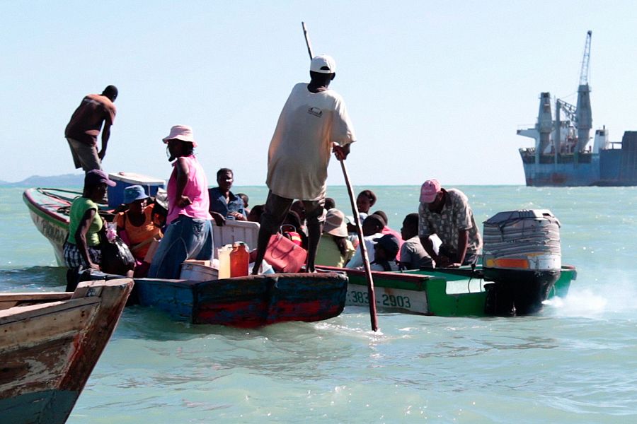 Barca de madera con numerosas personas navegando en aguas turquesa. Un hombre la impulsa con una vara.  Al fondo, un gran barco.