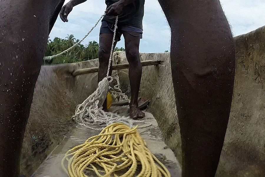 Hombre en canoa manipulando cuerdas y red de pesca. Flotadores amarillos y vegetación tropical de fondo.