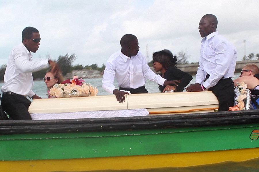 Funeral marítimo: ataúd blanco en barca verde y amarilla; personas vestidas de blanco lo transportan; mujer de luto en negro observa.