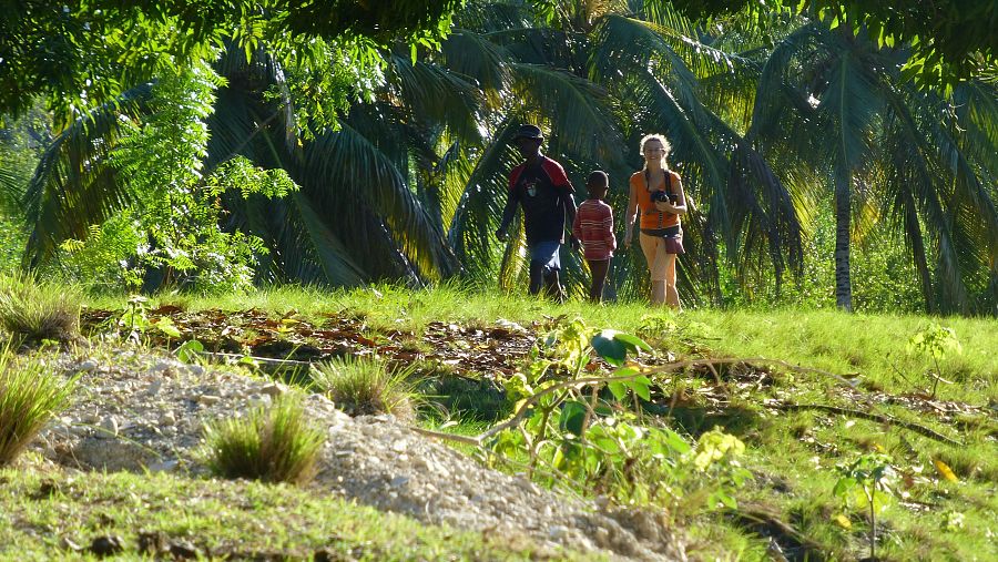 Personas caminando por un sendero tropical; una mujer con cámara, un hombre y un niño.  Vegetación exuberante con palmeras.