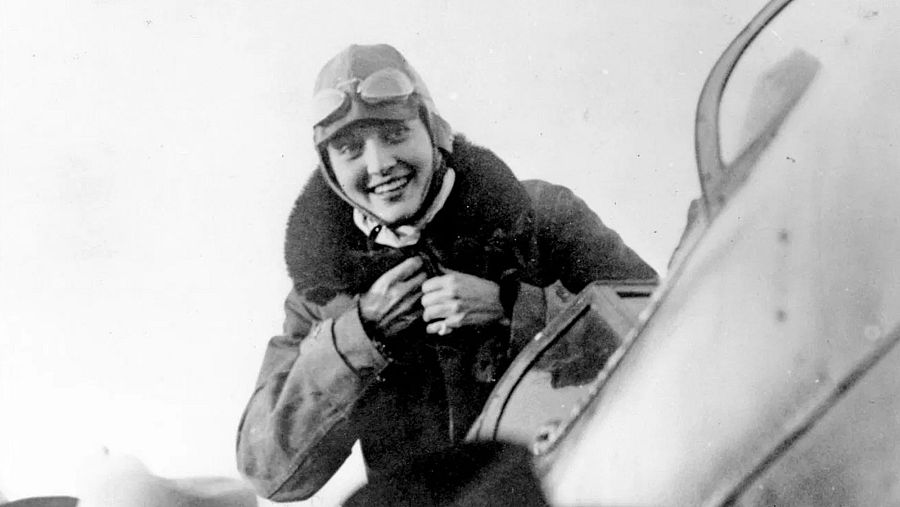 Aviadora con casco y gafas de cuero sonriendo junto a un avión. Imagen en blanco y negro, posiblemente de los años 20, que muestra confianza y determinación.