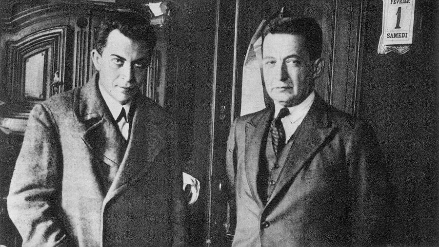 Dos hombres en traje, uno con abrigo, en foto antigua en blanco y negro. Calendario con fecha sábado 1 de febrero visible en la pared.