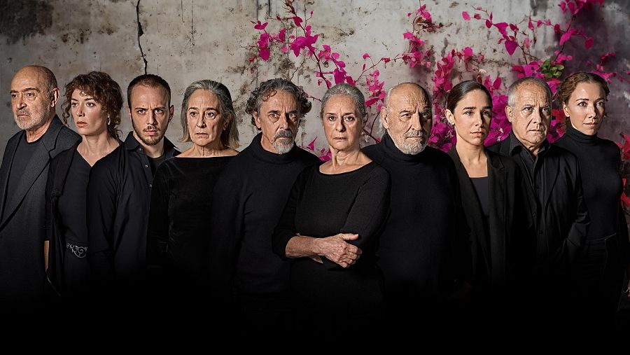 Diez actores, vestidos de negro, posan ante una pared agrietada con una buganvilla rosa. Fotografía del reparto completo de una obra de teatro.