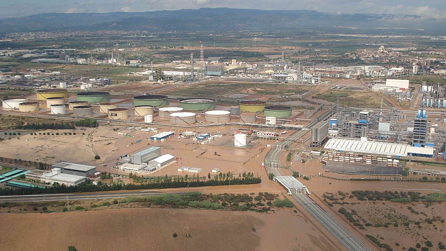 Inundación en planta química: tanques de almacenamiento cilíndricos parcialmente sumergidos en agua marrón.  Extensa área industrial afectada, incluyendo vía de comunicación parcialmente inundada.