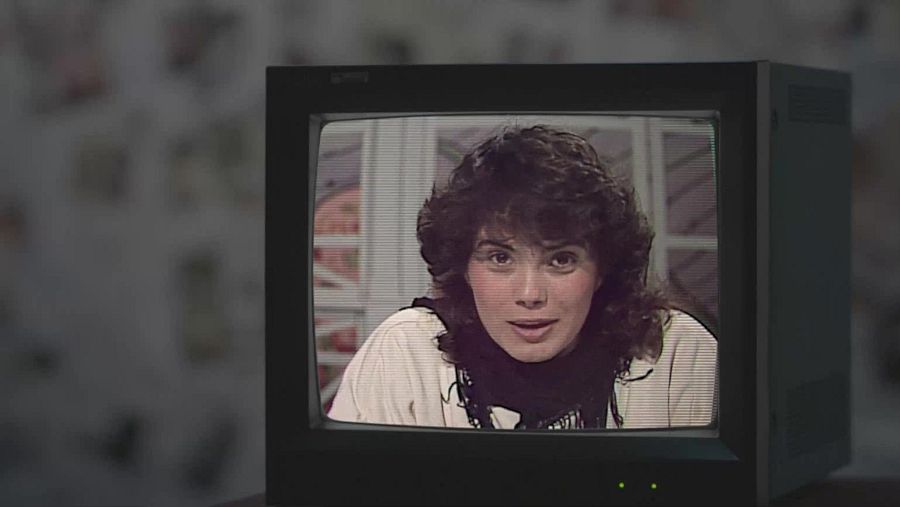 Televisor antiguo mostrando a una presentadora de televisión infantil de los años 80 con blusa clara y pañuelo oscuro, pelo corto y rizado. Fondo desenfocado.