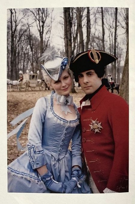 Dos figuras en trajes de época, ella con vestido azul y sombrero, él con traje rojo y tricornio. Fondo con carruaje y árboles sin hojas. Inspirado en el vestuario de la película de Sofía Coppola.