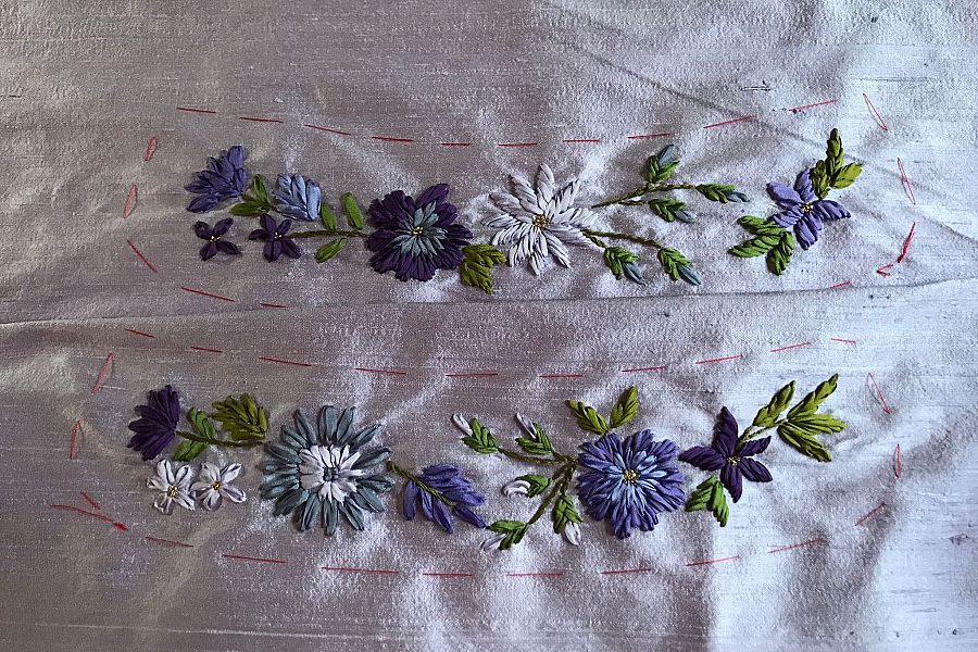 Bordado a mano con flores azules, violetas, blancas y verdes sobre tela gris. Hilos rojos marcan el diseño. Detalle de precisión y detalle en el trabajo.