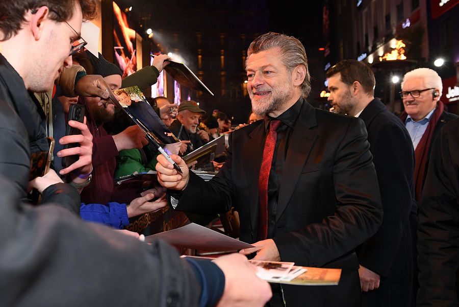 El actor Andy Serkis, con traje negro y corbata roja, firma autógrafos a fans en un evento público. Sonríe mientras interactúa con ellos.