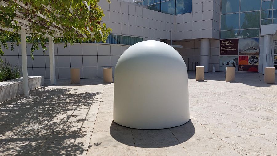 Escultura blanca, lisa y semiesférica en espacio abierto con bolardos y fachada de edificio moderno de fondo.