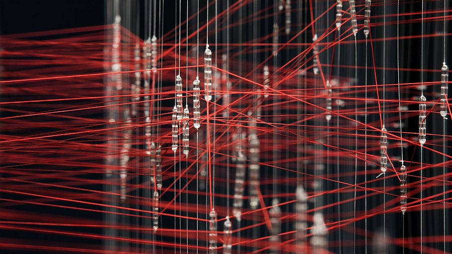 Instalación artística con hilos rojos y estructuras cilíndricas transparentes que cuelgan de una compleja red, creando una sensación de profundidad.