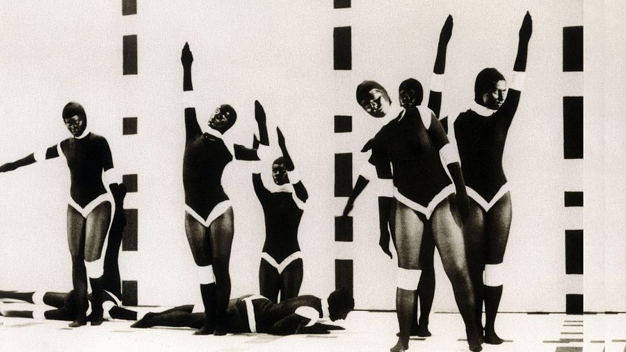 Imagen en blanco y negro de una pieza de danza contemporánea. Bailarines con trajes oscuros y detalles blancos en diferentes posturas sobre un fondo con franjas verticales. Movimiento y ritmo.