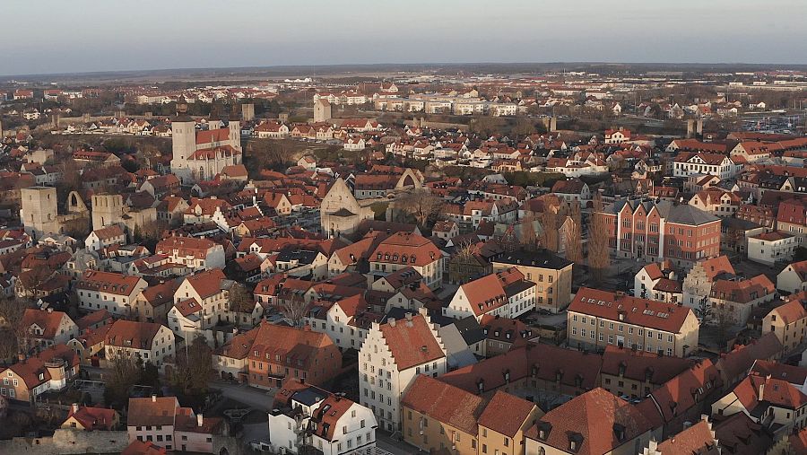 Vista aérea de una ciudad con numerosos tejados rojos, una gran estructura central y áreas verdes. Iluminación suave, posiblemente amanecer o atardecer.