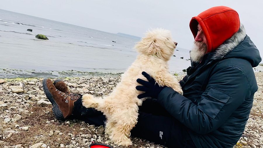 Hombre con capucha roja y botas marrones sentado en rocas junto a su perro, que está de pie sobre sus patas traseras.  Se ve agua al fondo y un frisbee rojo a sus pies.
