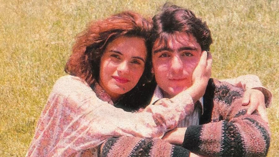 Pareja abrazada en un campo de hierba. Ella viste blusa floral, él camisa a rayas.  Imagen con posible relación a un documental.