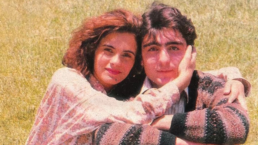 Pareja abrazada en un campo.  Ella lleva blusa rosa floral, él jersey a rayas.  Imagen cálida y cercana.