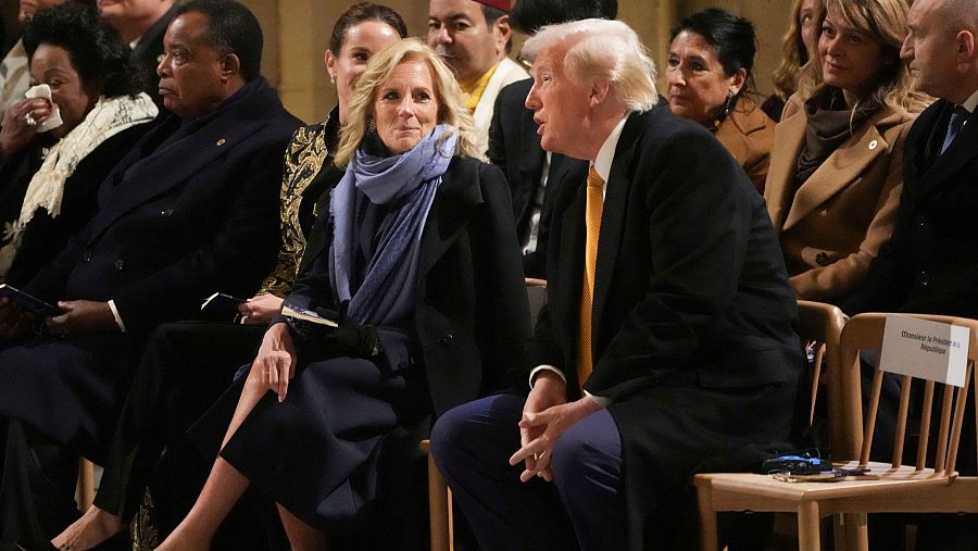 Trump y Biden conversan sentados en un evento formal. Él viste traje oscuro y corbata dorada; ella, abrigo oscuro y bufanda azul. Una mujer llora cerca.