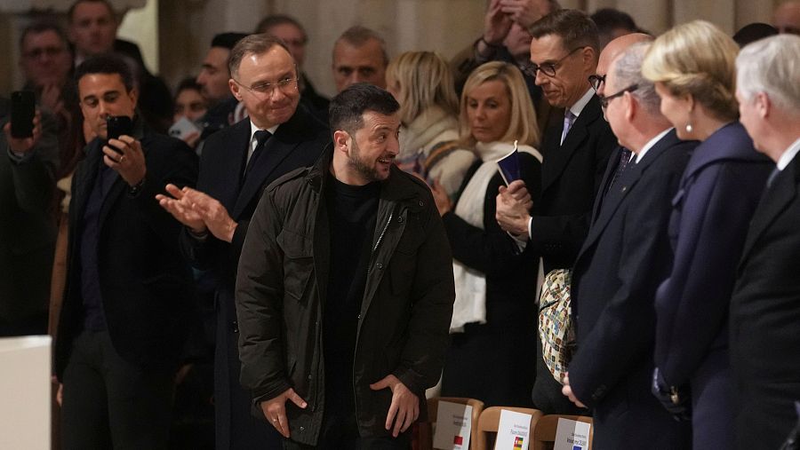 Volodímir Zelenski, con chaqueta oscura, saluda a invitados en la catedral de Notre Dame.  Personas en ropa formal lo rodean; algunos aplauden.